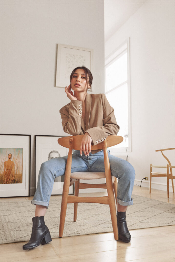 Nadya Inzunza is shown seated on a wooden chair in a minimalist room with framed artworks on the wall. She wears a beige blazer blue jeans and black ankle boots.